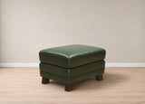 A square green upholstered ottoman with dark brown wooden legs, shown in a minimalist room setting with a beige textured wall and light wood floor.