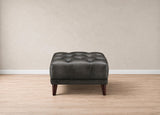 A dark gray tufted leather ottoman with dark wooden legs, positioned against a neutral beige wall and light wood floor.