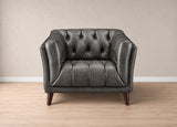 A dark gray tufted leather armchair with dark wooden legs, positioned against a neutral beige wall and light wood floor.