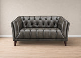 A dark gray tufted leather loveseat with dark wooden legs, set against a neutral beige wall and light wood floor.