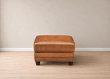A square brown leather ottoman with dark wooden legs and nailhead trim, centered against a neutral beige wall and light wood floor.
