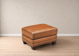 A square brown leather ottoman with dark wooden legs and nailhead trim, set against a neutral beige wall and light wood floor.