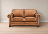 A classic two-seater loveseat sofa upholstered in warm tan leather, featuring rolled arms with nailhead trim, dark wooden legs, and a clean studio setting with a neutral beige wall and light wood floor.