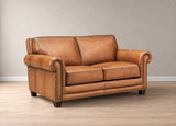 A classic two-seater loveseat sofa upholstered in warm tan leather with rolled arms, nailhead trim, and dark wooden legs, set against a neutral beige wall and light wood floor.