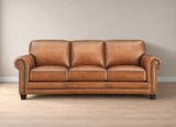 A classic three-seater leather sofa in a warm tan/cognac brown, featuring rolled arms with nailhead trim, deep seat cushions, and dark wooden legs, set against a neutral beige wall and light wood floor.