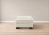 A square upholstered ottoman in a light cream fabric with dark brown wooden legs, set against a neutral beige wall and light wood floor.
