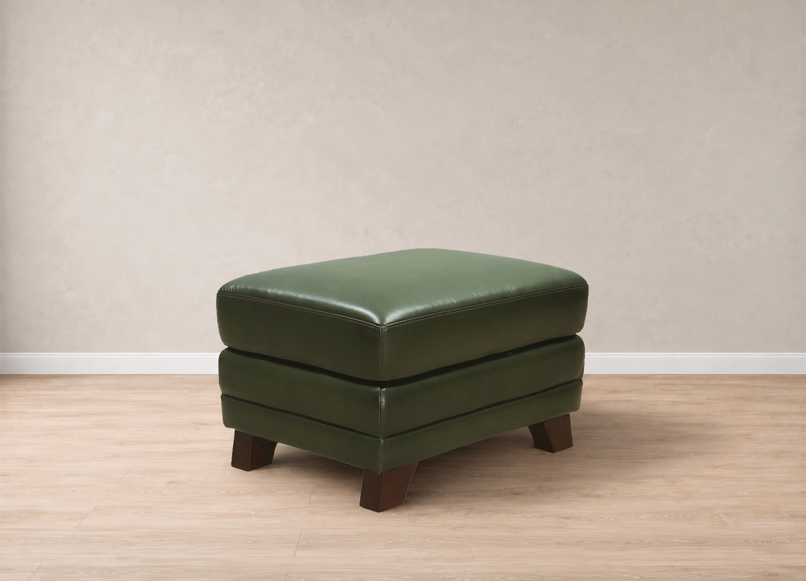 A square green upholstered ottoman with dark brown wooden legs, shown in a minimalist room setting with a beige textured wall and light wood floor.