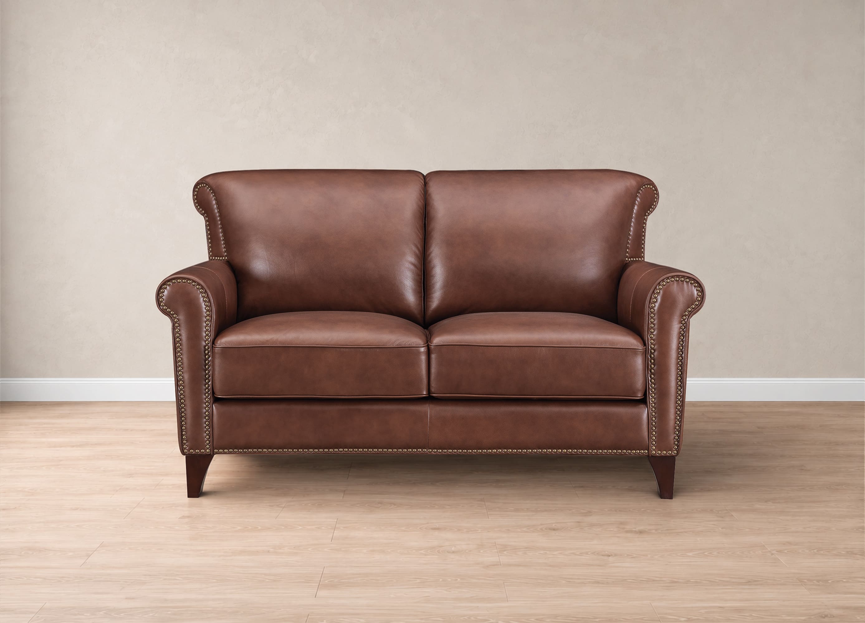 A classic brown leather loveseat sofa with rolled arms, nailhead trim, and dark wooden legs, set against a neutral beige wall and light wood floor.