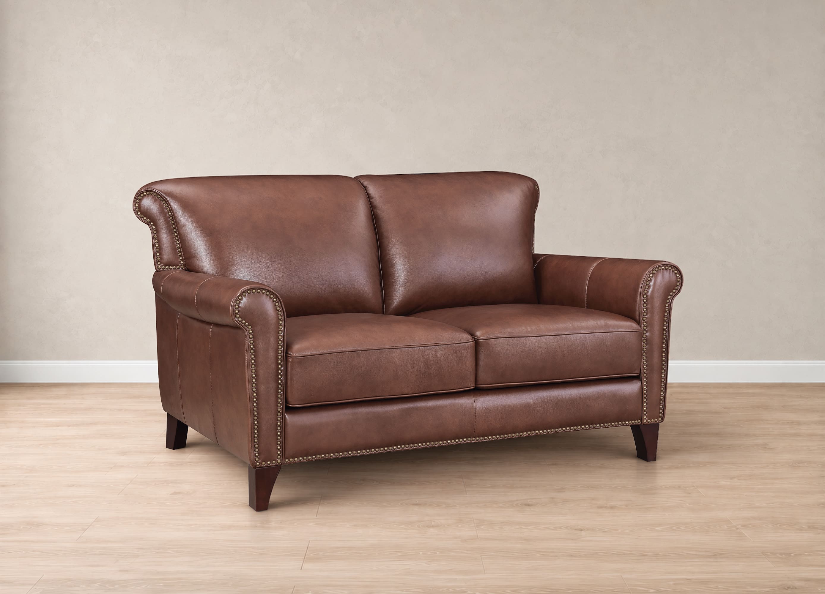 A classic two-seater loveseat sofa upholstered in rich brown leather, featuring rolled arms, nailhead trim, and dark wooden legs, set against a neutral beige wall and light wood floor.
