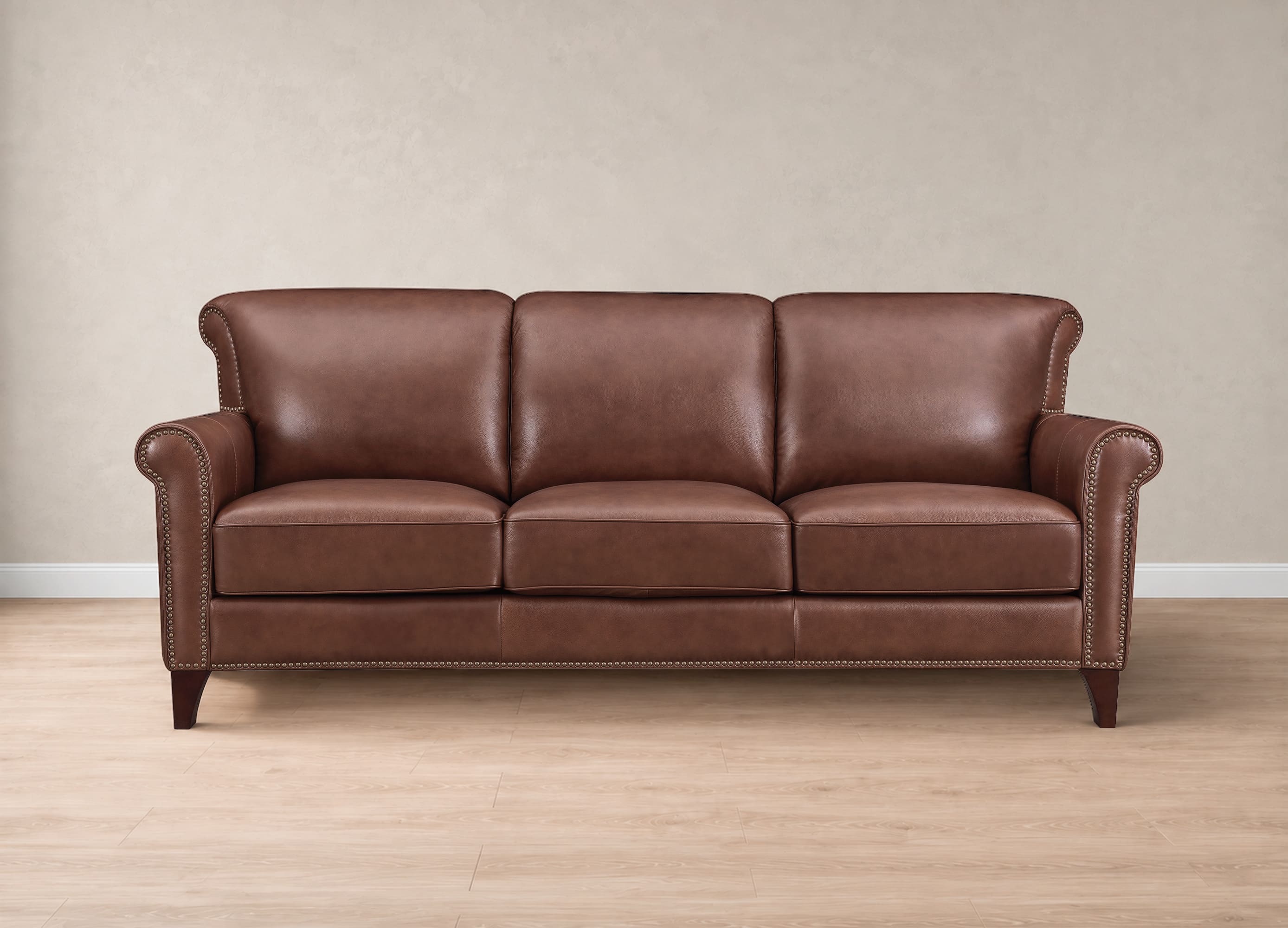 A classic three-seater brown leather sofa with rolled arms, nailhead trim, and dark wooden legs, set against a neutral beige wall and light wood floor.