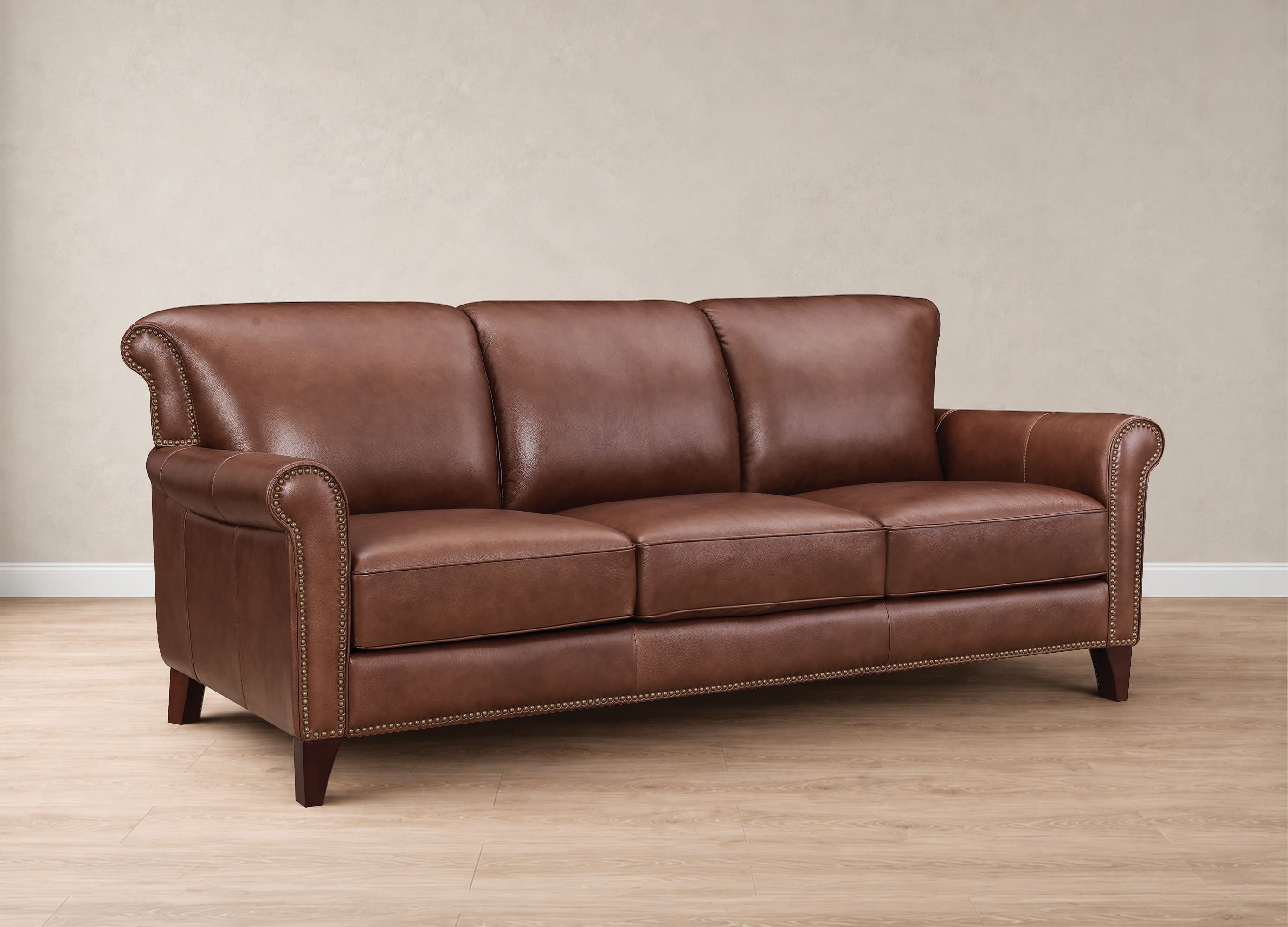A classic three-seater leather sofa in a warm brown color, featuring rolled arms, nailhead trim, and dark wooden legs, set against a neutral beige wall and light wood floor.
