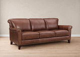 A classic three-seater leather sofa in a warm brown color, featuring rolled arms, nailhead trim, and dark wooden legs, set against a neutral beige wall and light wood floor.