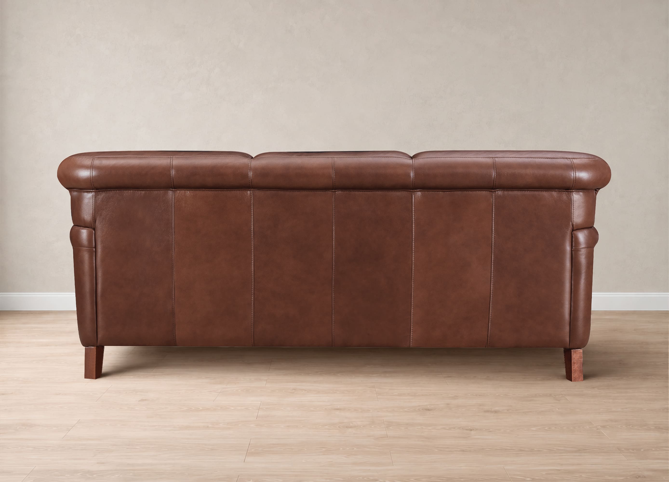 A brown leather sofa with a tufted backrest and rolled arms, shown from the rear against a neutral beige wall and light wood floor.