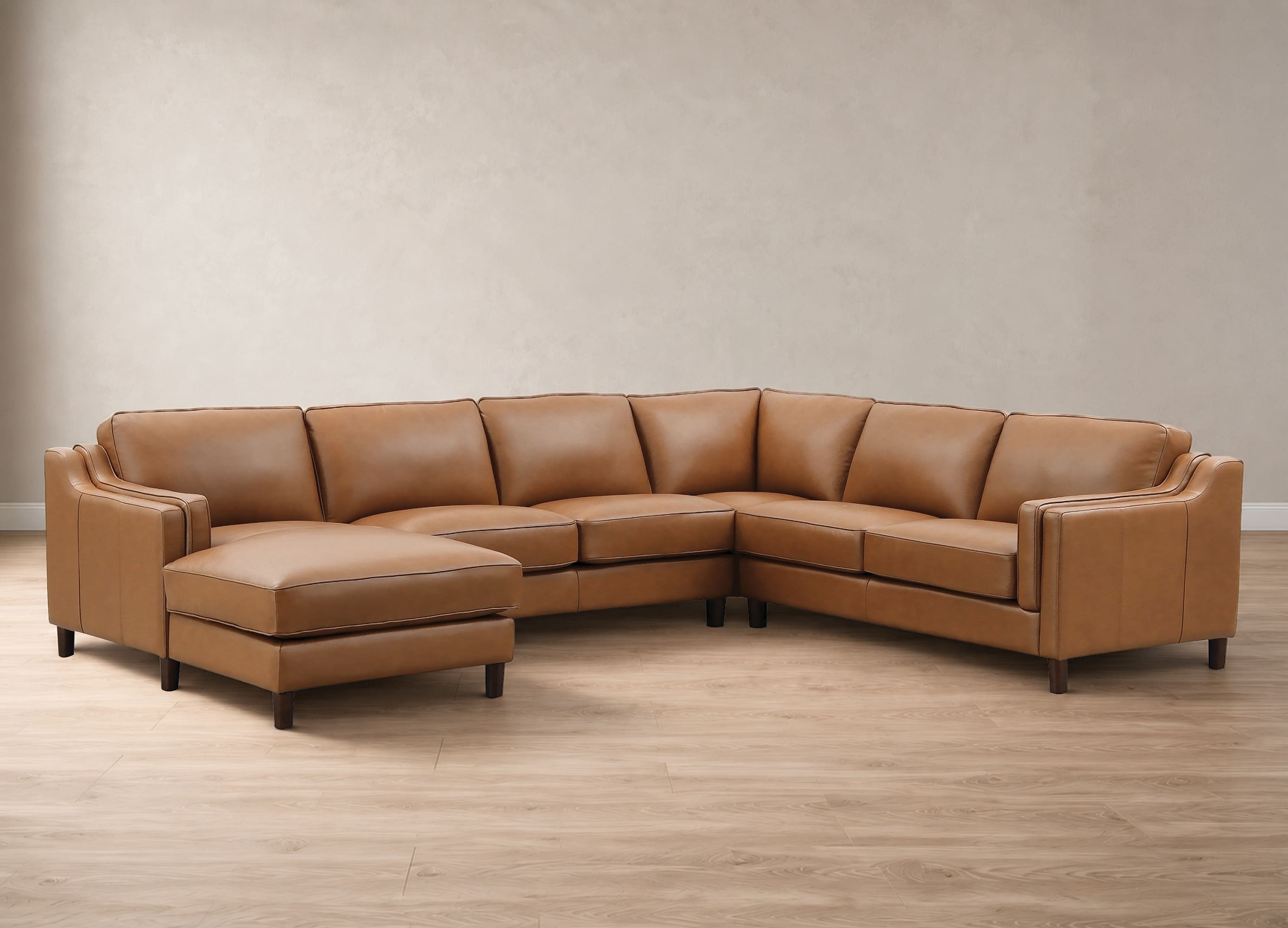A curved sectional sofa upholstered in warm tan leather, featuring a left-facing chaise ottoman and dark wooden legs, set against a neutral beige wall and light wood floor.