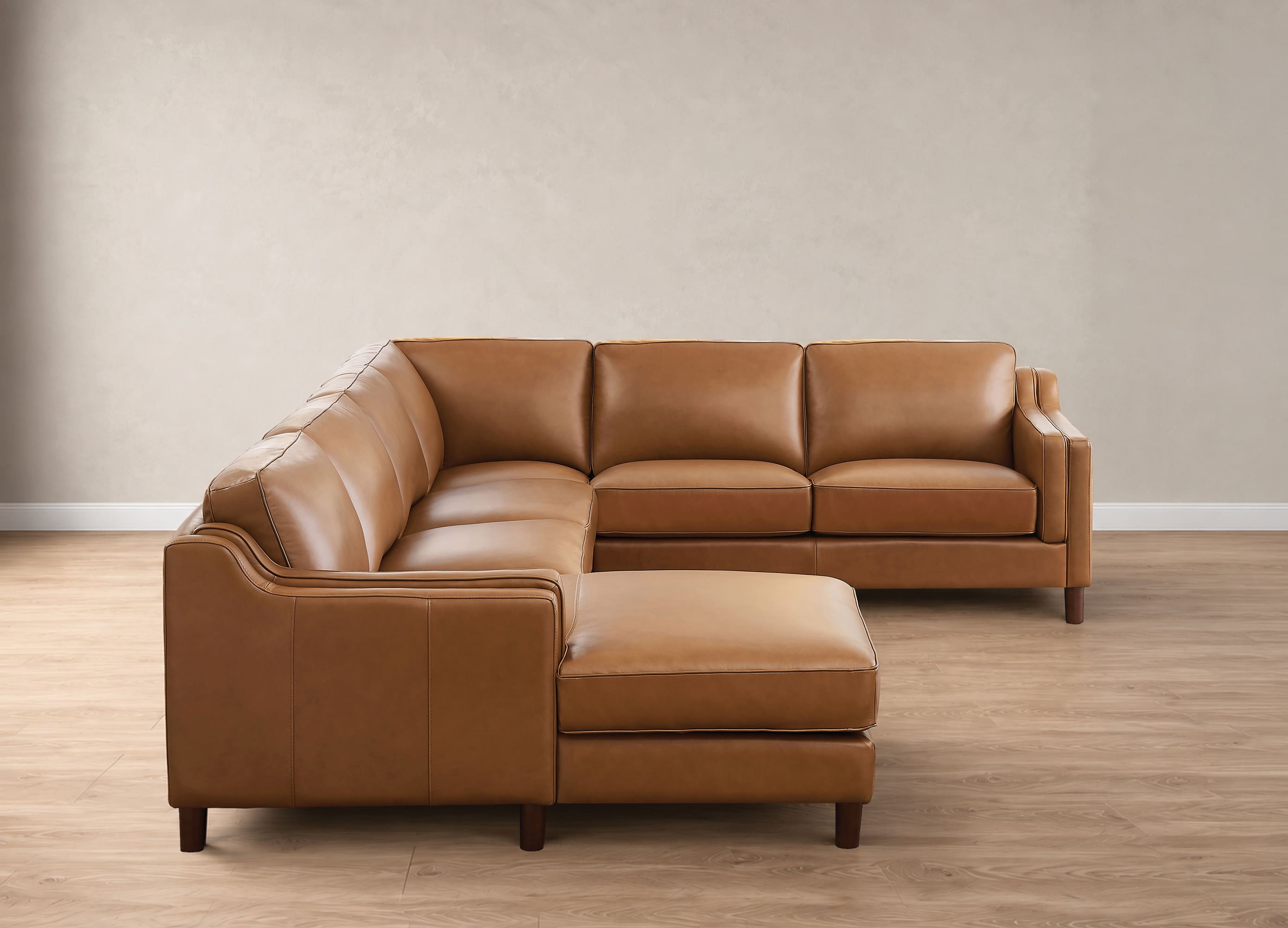 A modern L-shaped sectional sofa in tan leather with a matching chaise ottoman, set against a neutral beige wall and light wood floor.