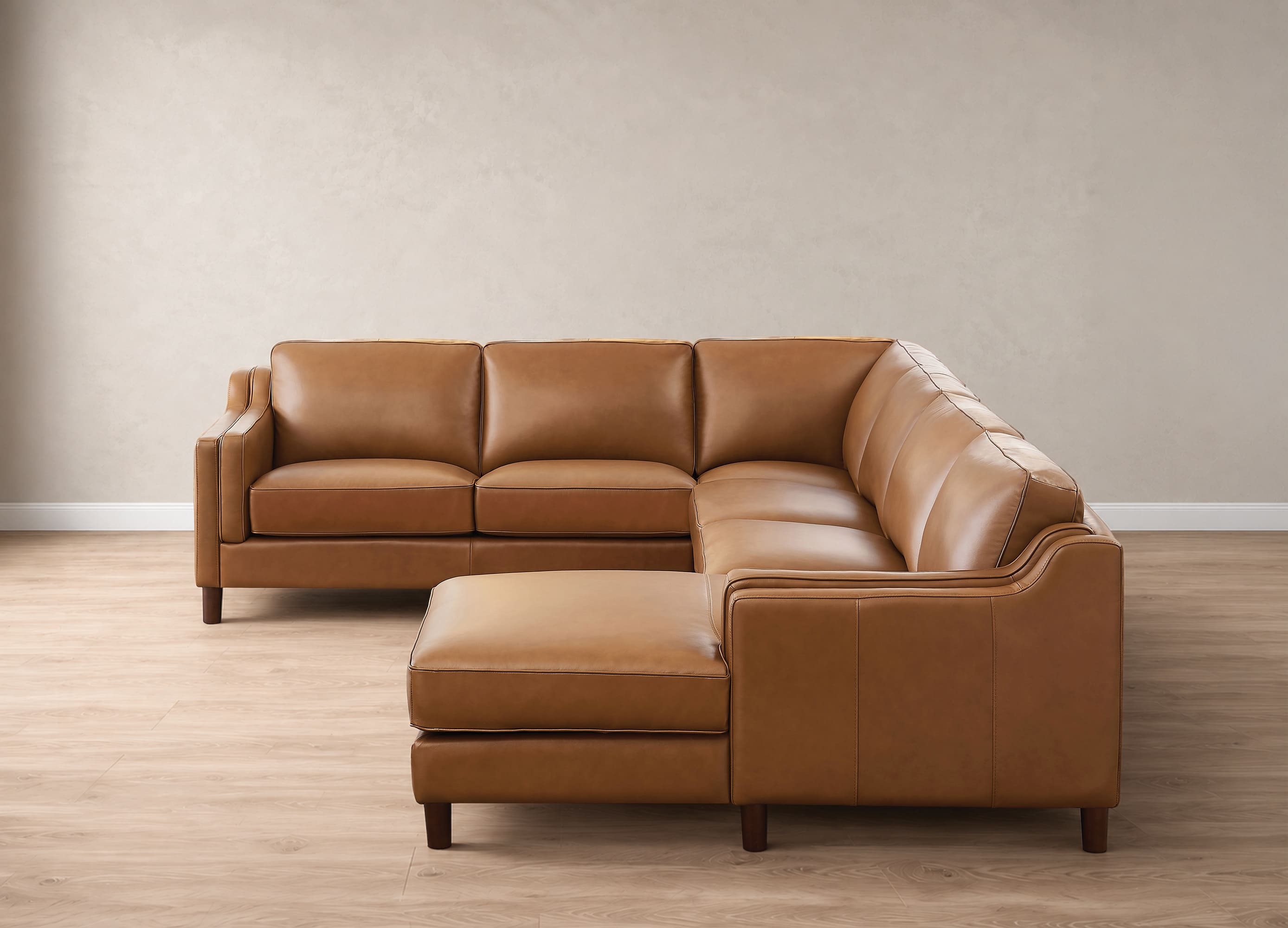 A modern sectional sofa with a chaise ottoman in tan leather, set against a neutral beige wall and light wood floor.