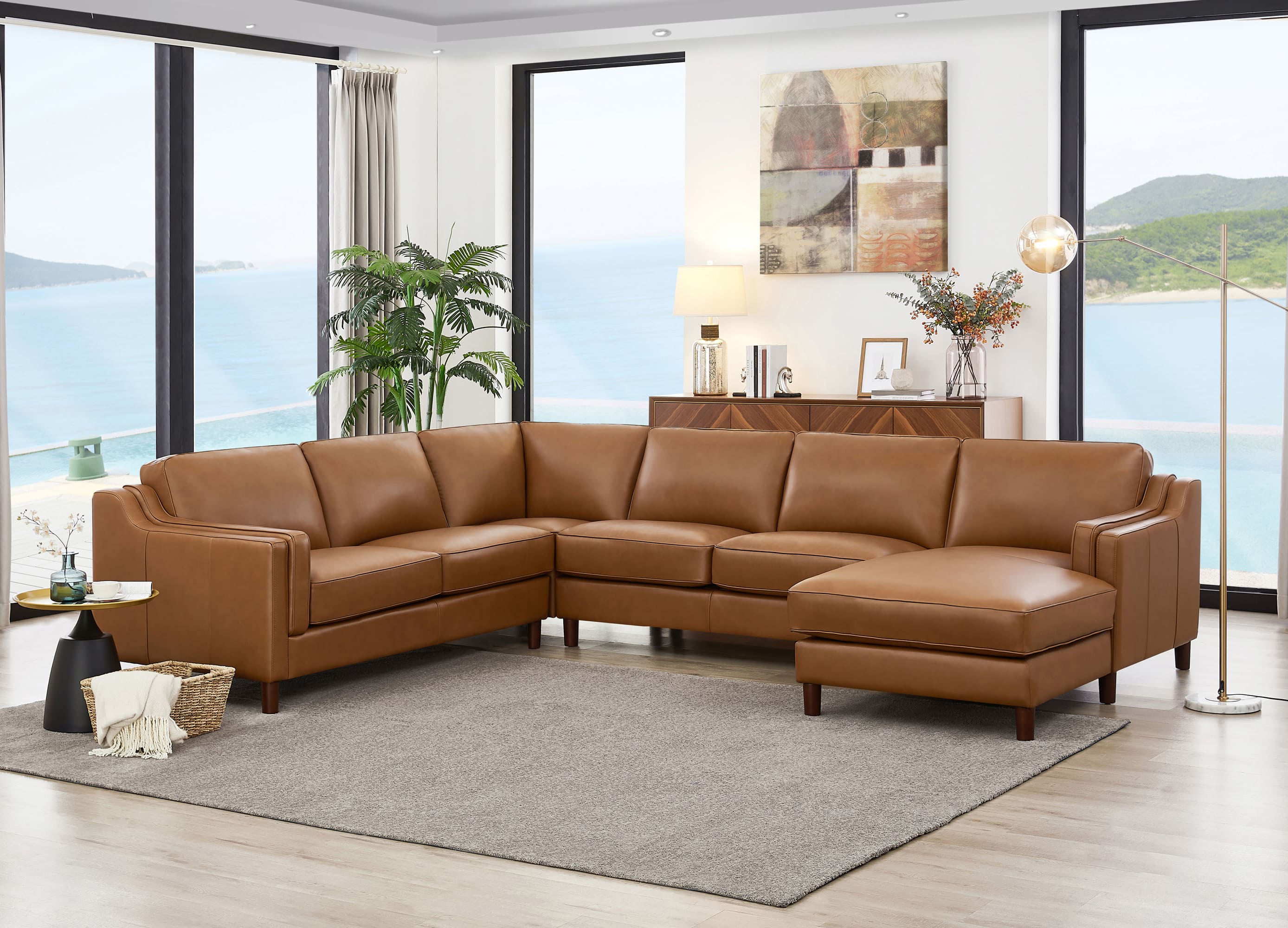 A tan leather sectional sofa with a chaise lounge, positioned on a gray area rug in a modern living room with large windows overlooking the ocean.