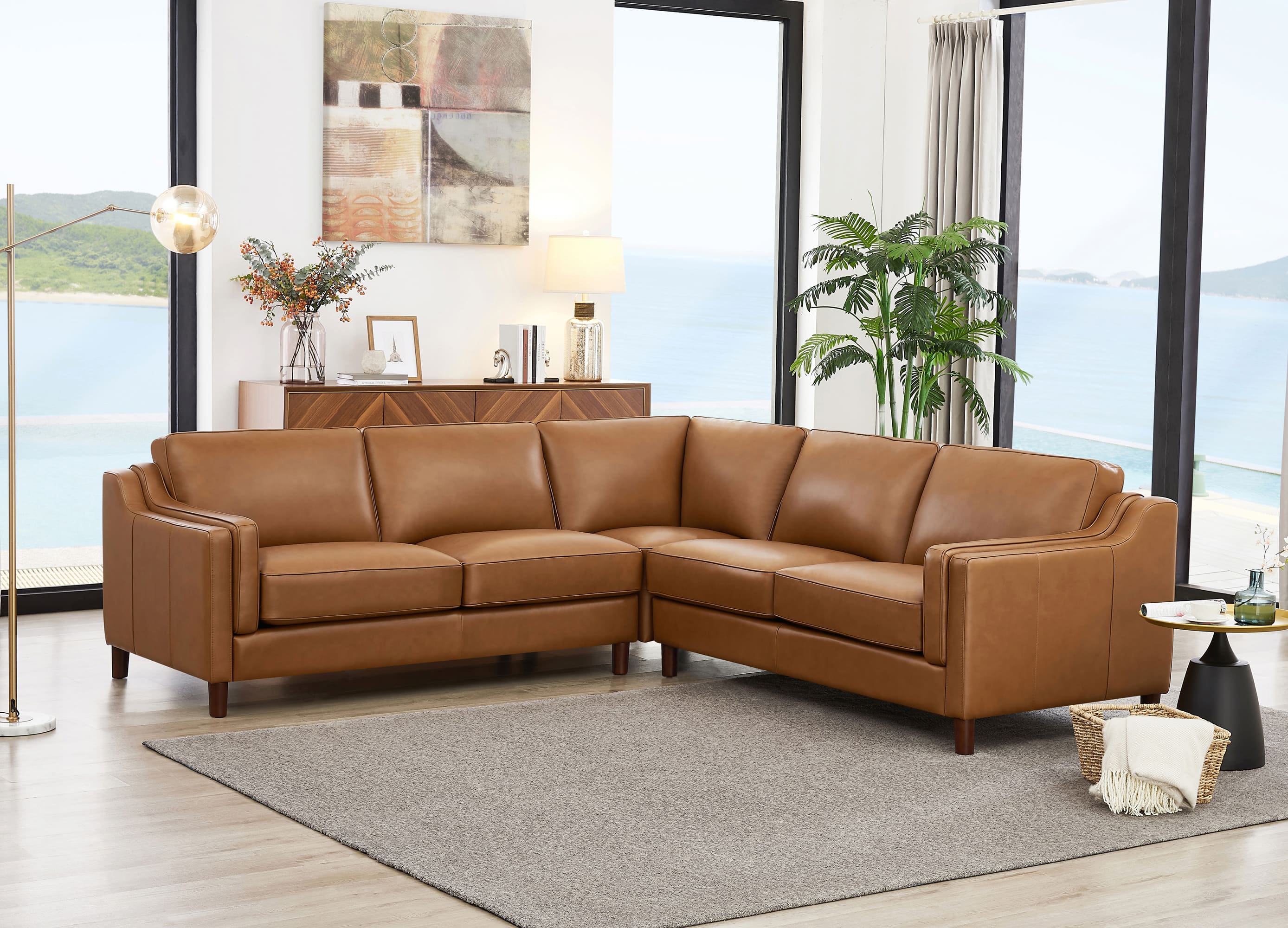 A modern L-shaped sectional sofa upholstered in warm tan leather, positioned in a bright, contemporary living room with large windows overlooking the water.
