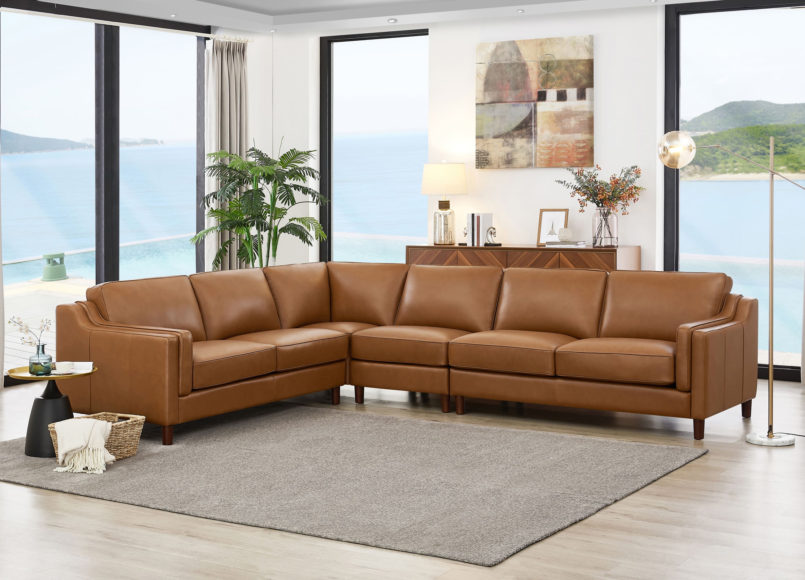 A modern L-shaped sectional sofa upholstered in warm tan leather, positioned on a large gray area rug in a bright, contemporary living room with large windows overlooking the ocean.