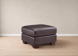 A dark brown upholstered ottoman with a square cushioned top and short wooden legs, shown in a minimalist room setting with light wood flooring and a beige textured wall.
