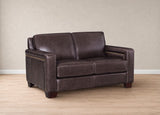 A dark brown leather loveseat sofa with nailhead trim on the arms, shown in a room setting with a beige wall and light wood floor.
