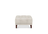 A square tufted ottoman in a light beige fabric, supported by four dark brown wooden legs, shown against a plain white background.
