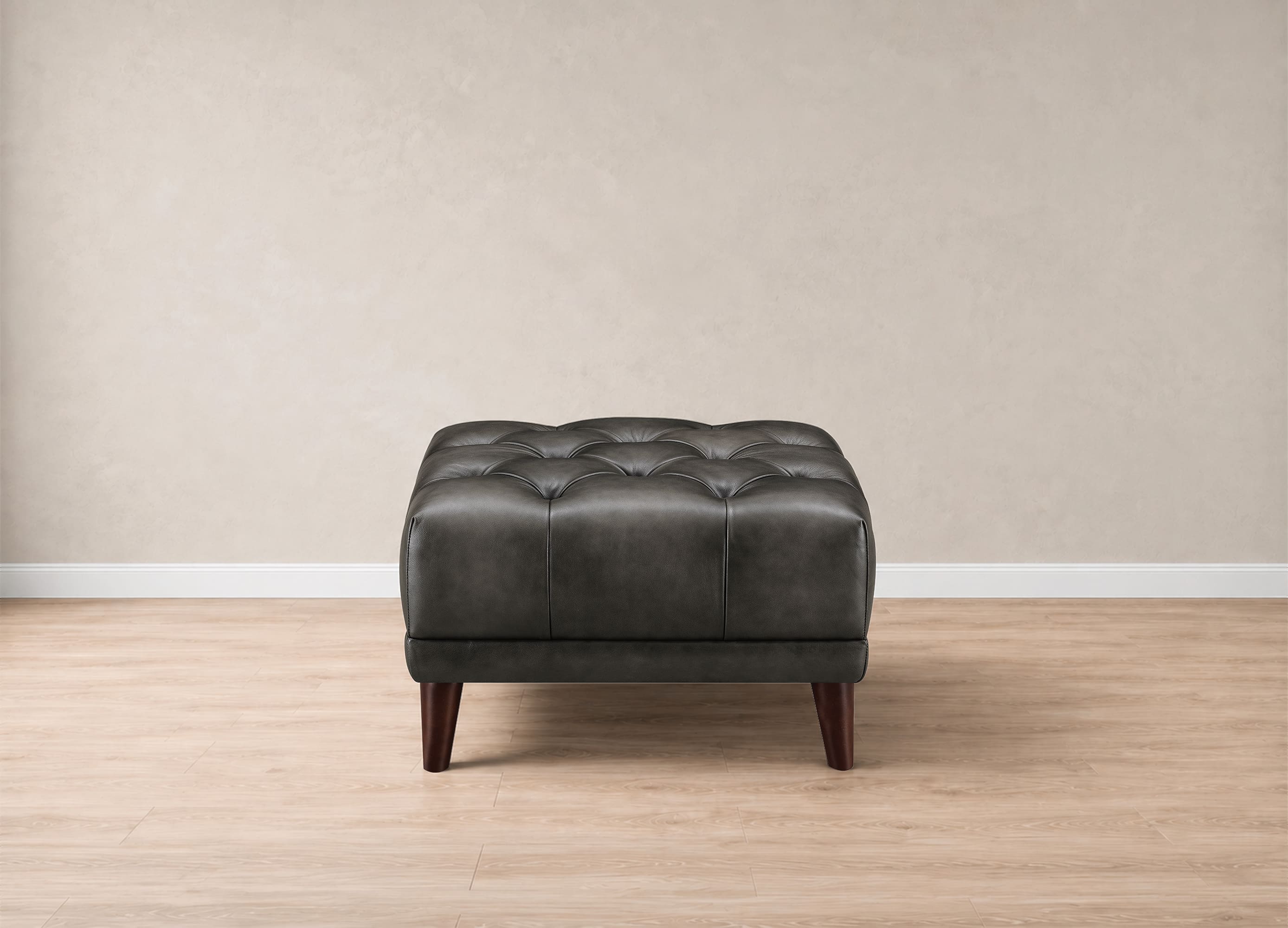 A dark gray tufted leather ottoman with dark wooden legs, positioned against a neutral beige wall and light wood floor.