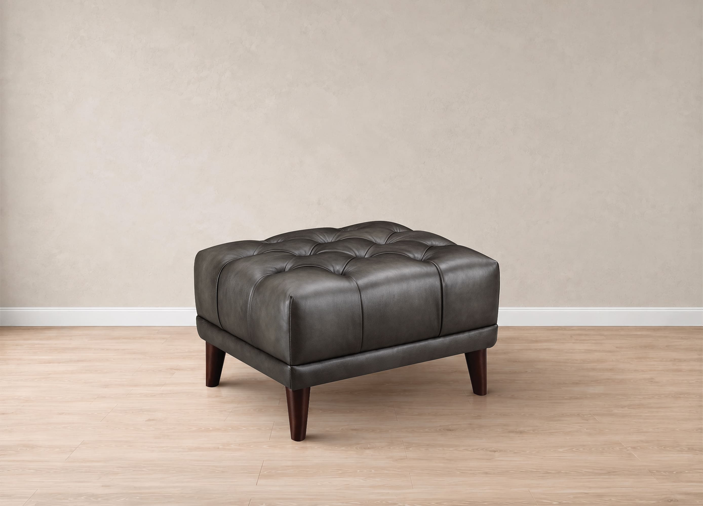 A square tufted ottoman in dark charcoal gray faux leather with dark brown wooden legs, set against a neutral beige wall and light wood floor.