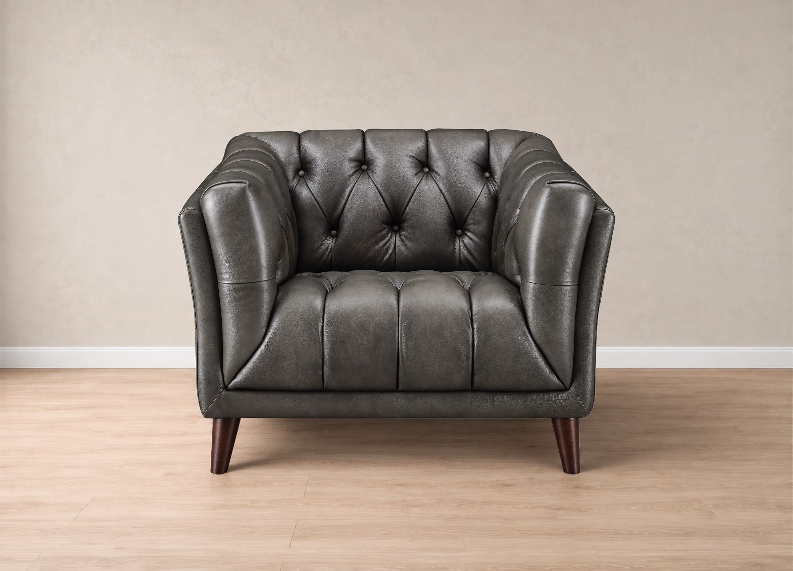 A dark gray tufted leather armchair with dark wooden legs, positioned against a neutral beige wall and light wood floor.