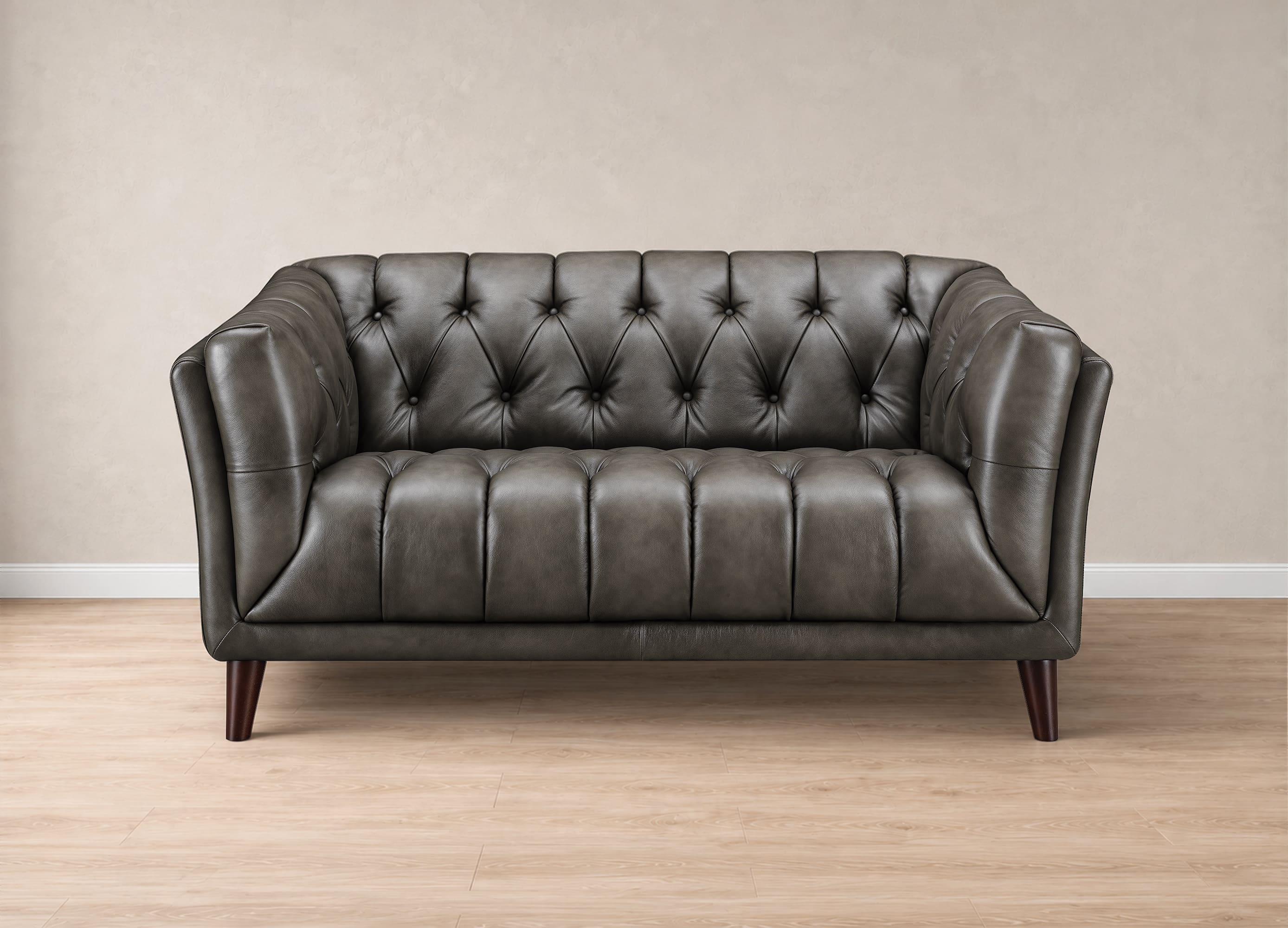 A dark gray tufted leather loveseat with dark wooden legs, set against a neutral beige wall and light wood floor.