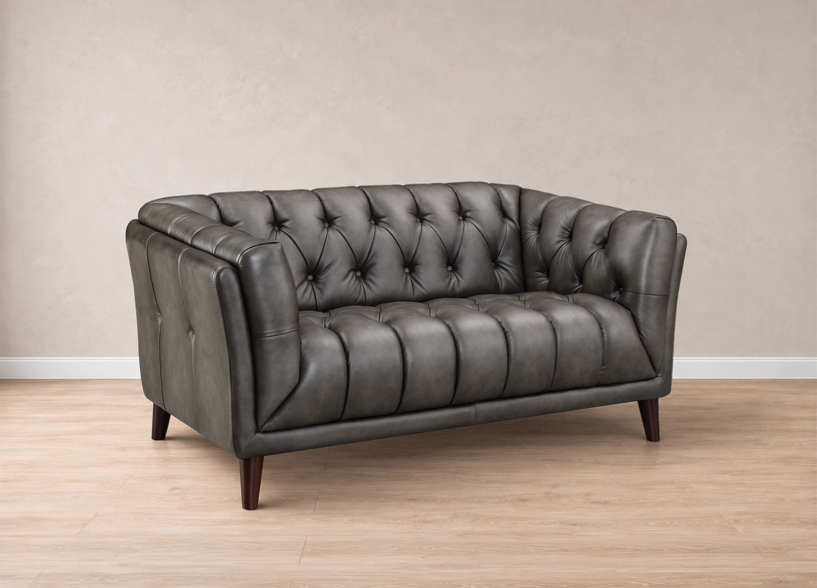 A dark gray tufted leather loveseat with dark wooden legs, positioned on a light wood floor against a neutral beige wall.