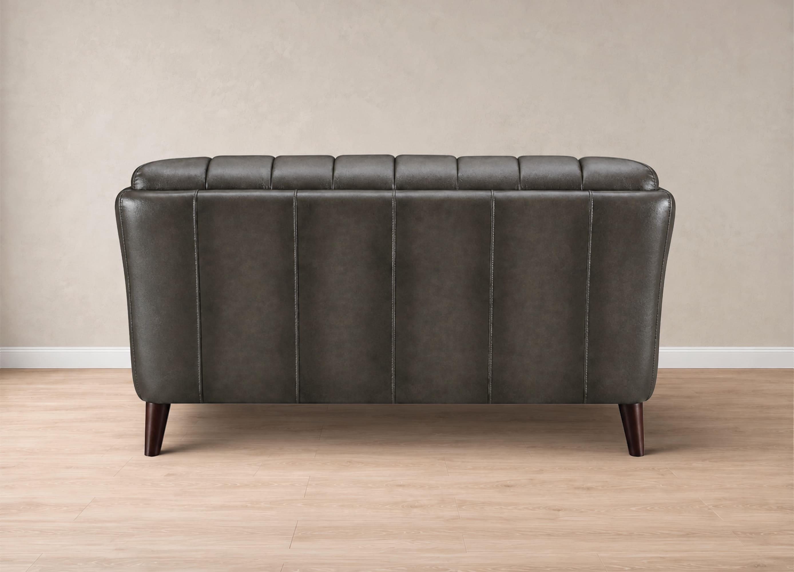 A dark gray tufted faux leather loveseat with dark wooden legs, shown from the back in a minimalist room setting with a beige wall and light wood floor.