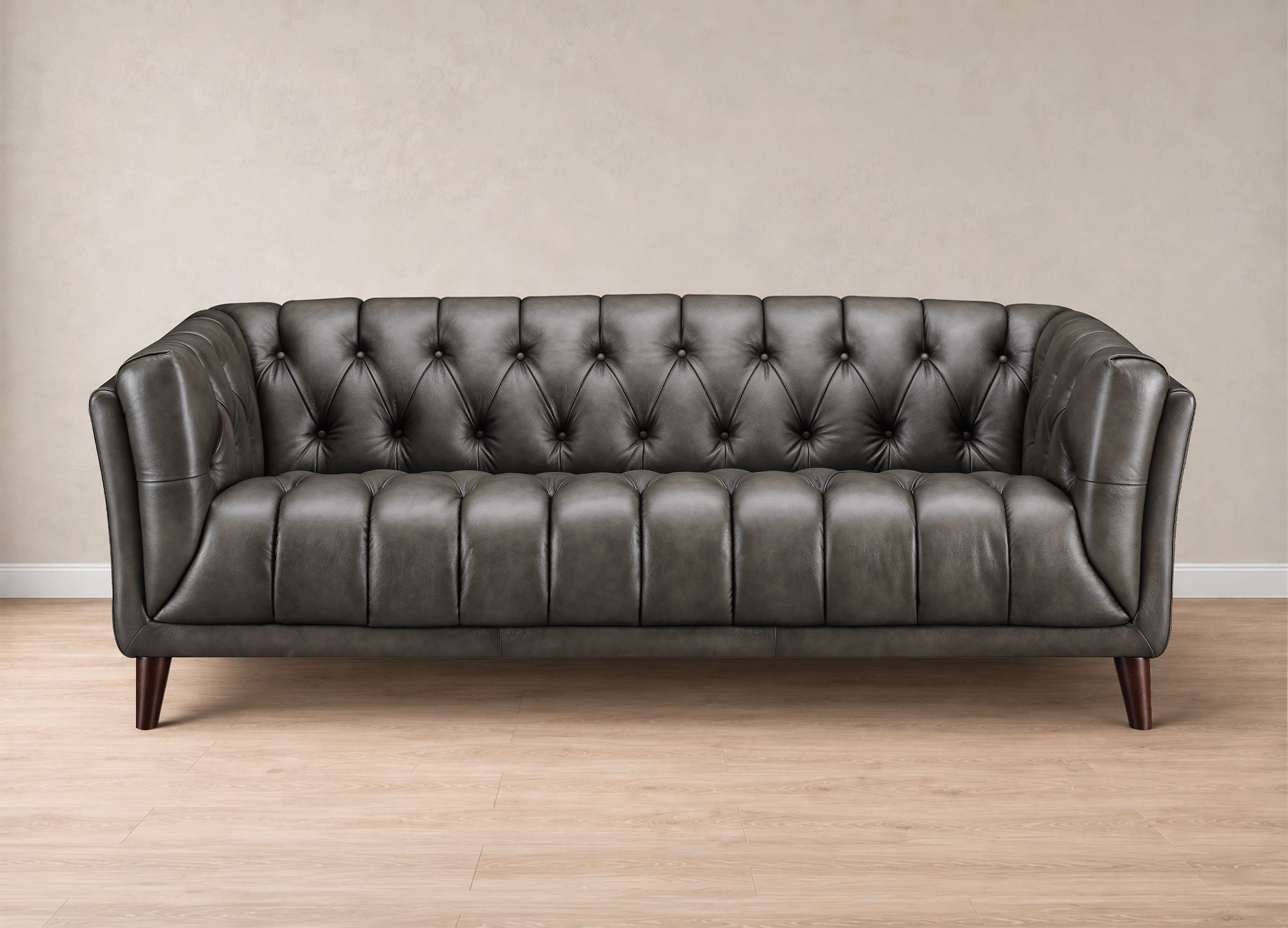 A dark gray tufted leather sofa with dark wooden legs, set against a neutral beige wall and light wood floor.