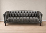 A dark gray tufted leather sofa with dark wooden legs, set against a neutral beige wall and light wood floor.