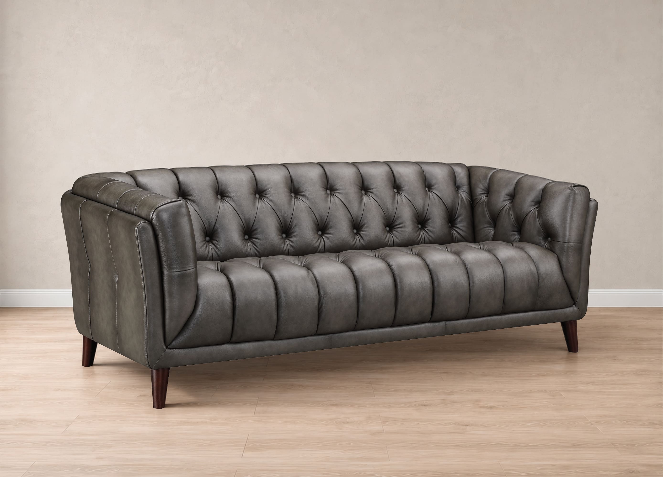 A dark gray tufted leather loveseat sofa with dark wooden legs, set against a neutral beige wall and light wood floor.