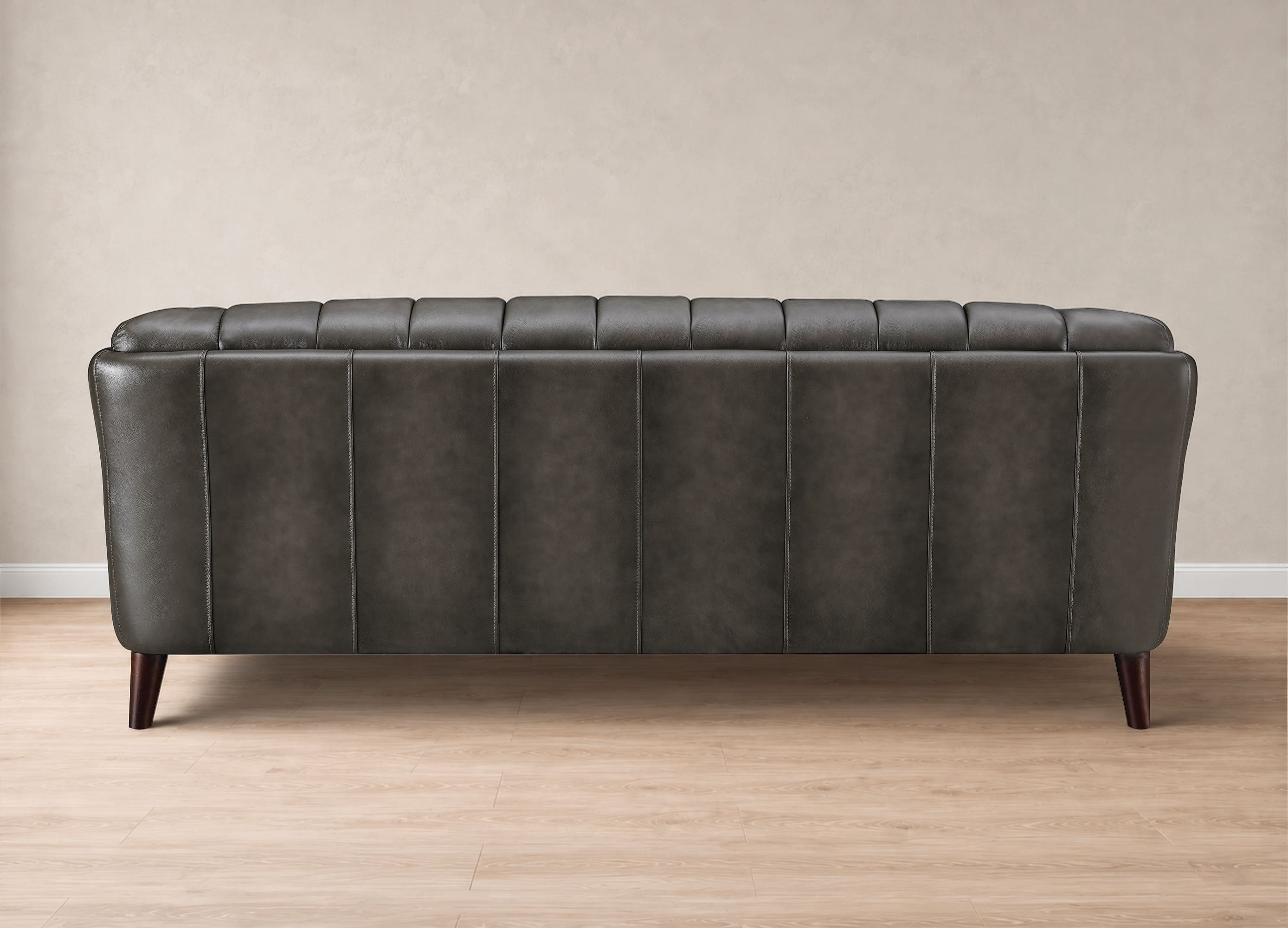 A dark gray tufted leather sofa shown from the back, featuring vertical channel tufting and dark wooden legs, set against a neutral beige wall and light wood floor.