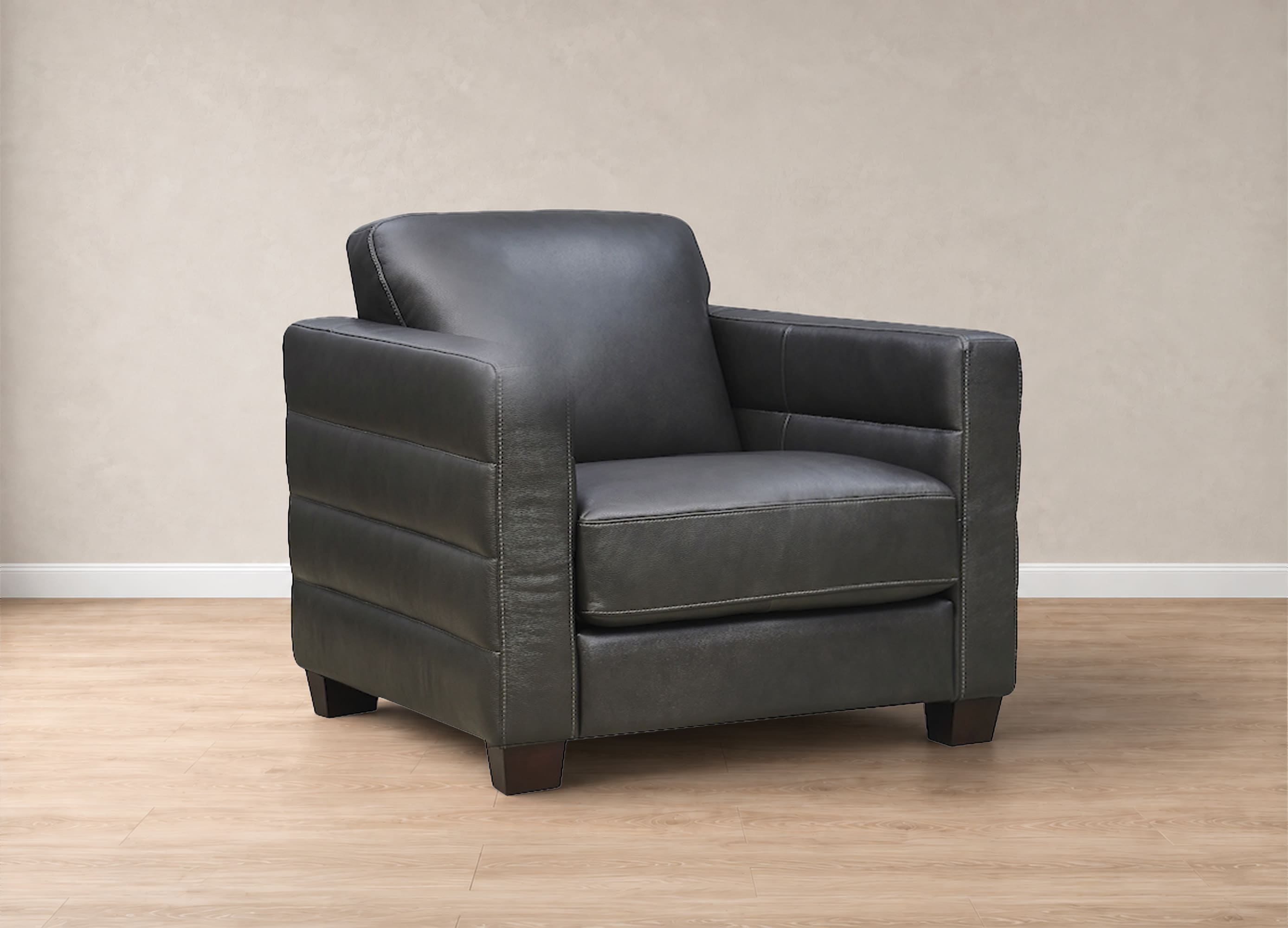 A dark gray leather armchair with a square backrest and horizontal channel tufting on the sides, shown on a light wood floor against a textured beige wall.