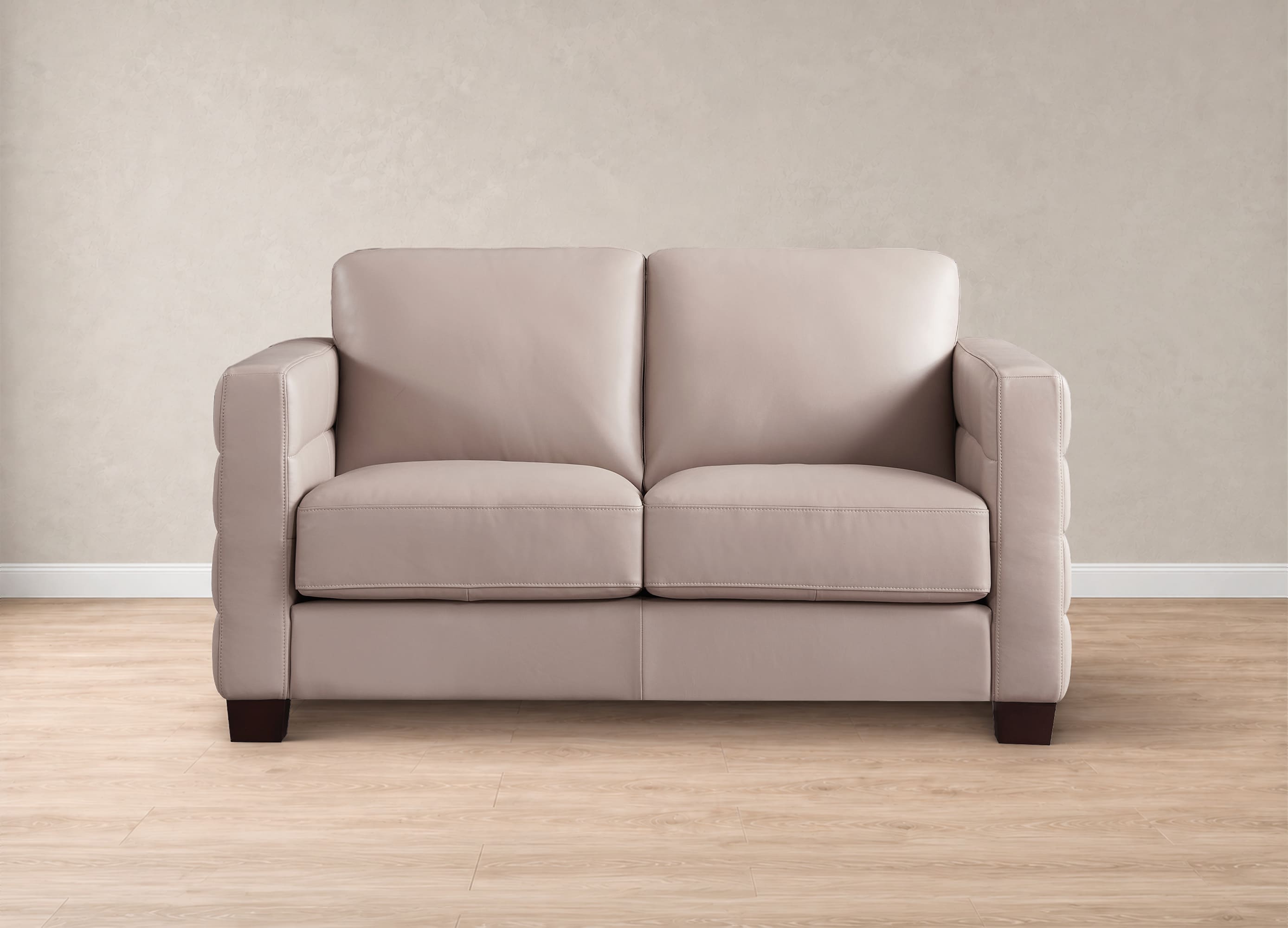 A two-seater loveseat sofa upholstered in a light beige fabric, positioned against a textured beige wall on a light wood floor.