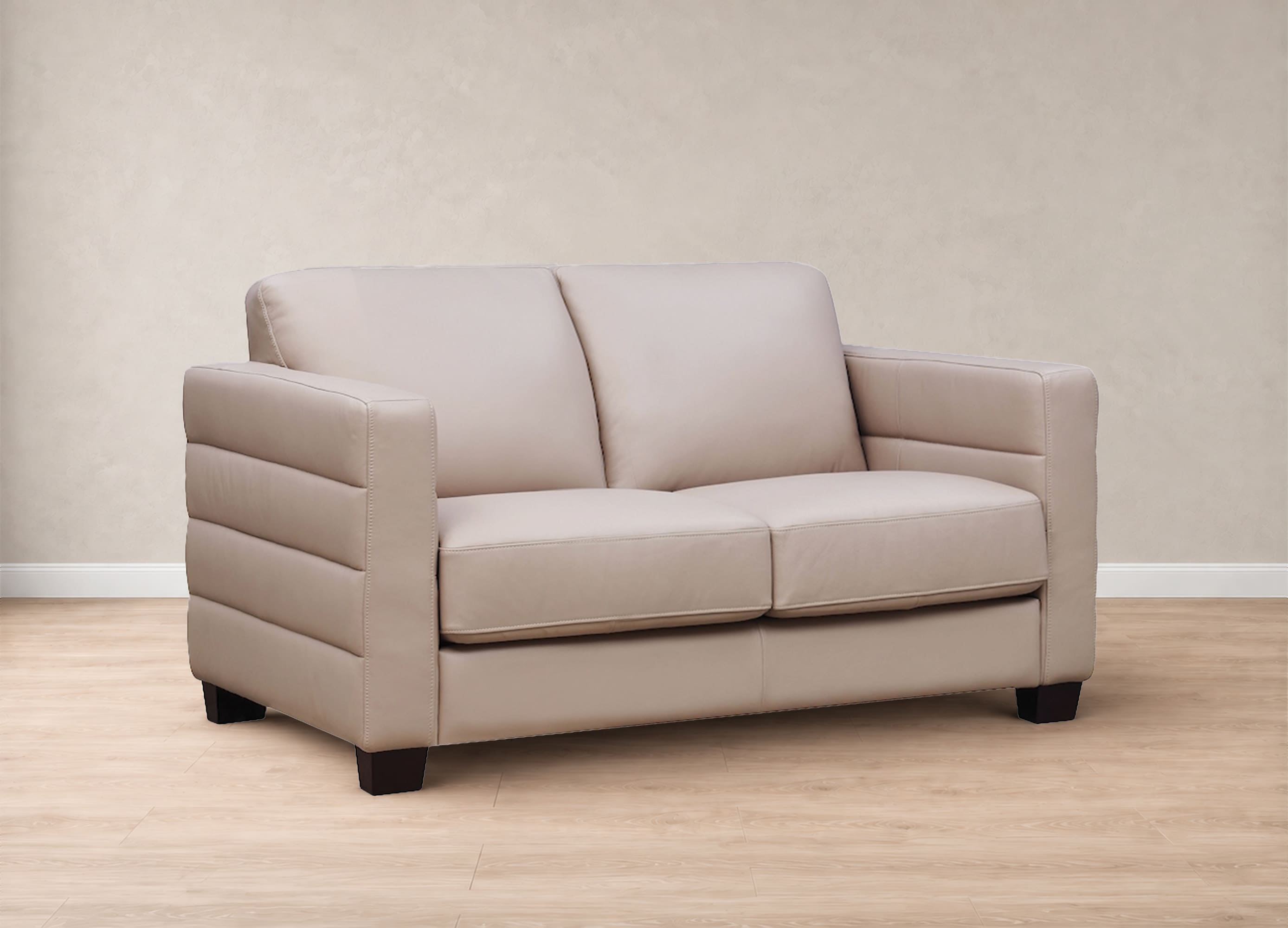 A two-seater loveseat sofa upholstered in light beige leather, featuring horizontal channel tufting on the arms and dark wooden legs, set against a neutral beige textured wall and light wood floor.