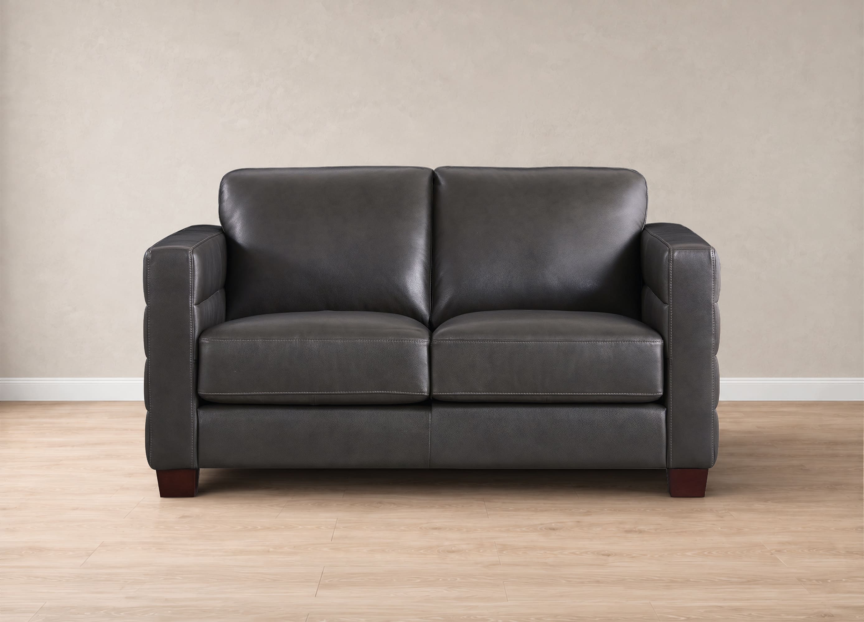 A dark gray leather loveseat sofa with square arms and dark wooden legs, shown from a straight-on perspective against a beige textured wall and light wood floor.