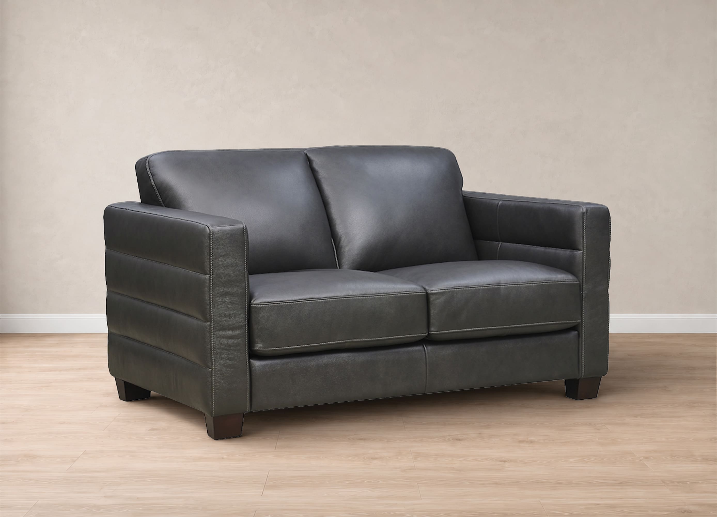A dark gray leather loveseat sofa with a modern design, featuring horizontal channel tufting on the arms and backrest, set against a neutral beige wall and light wood flooring.