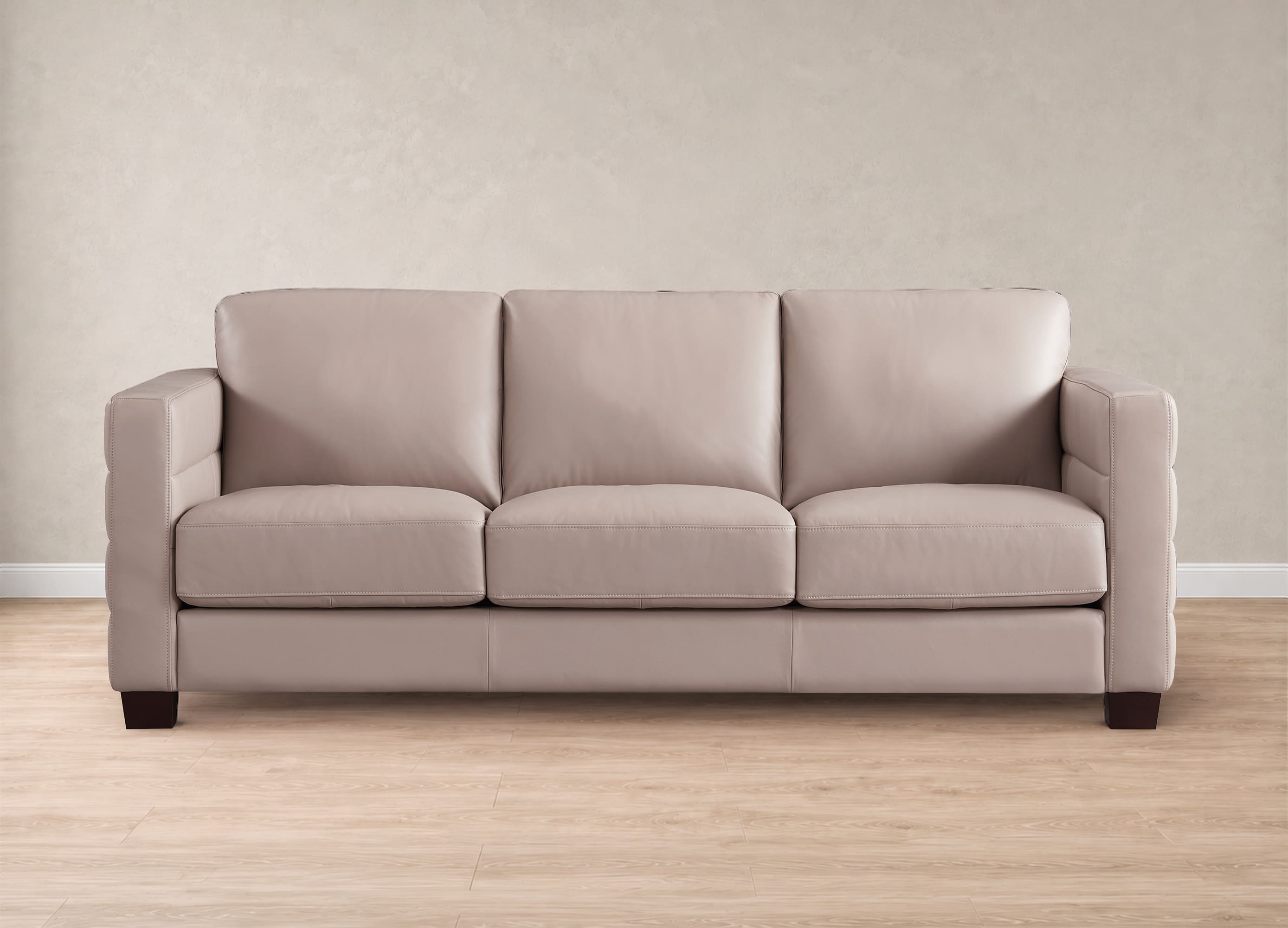 A three-seat upholstered sofa in a light beige or taupe fabric, shown from the front against a neutral beige wall and light wood floor.