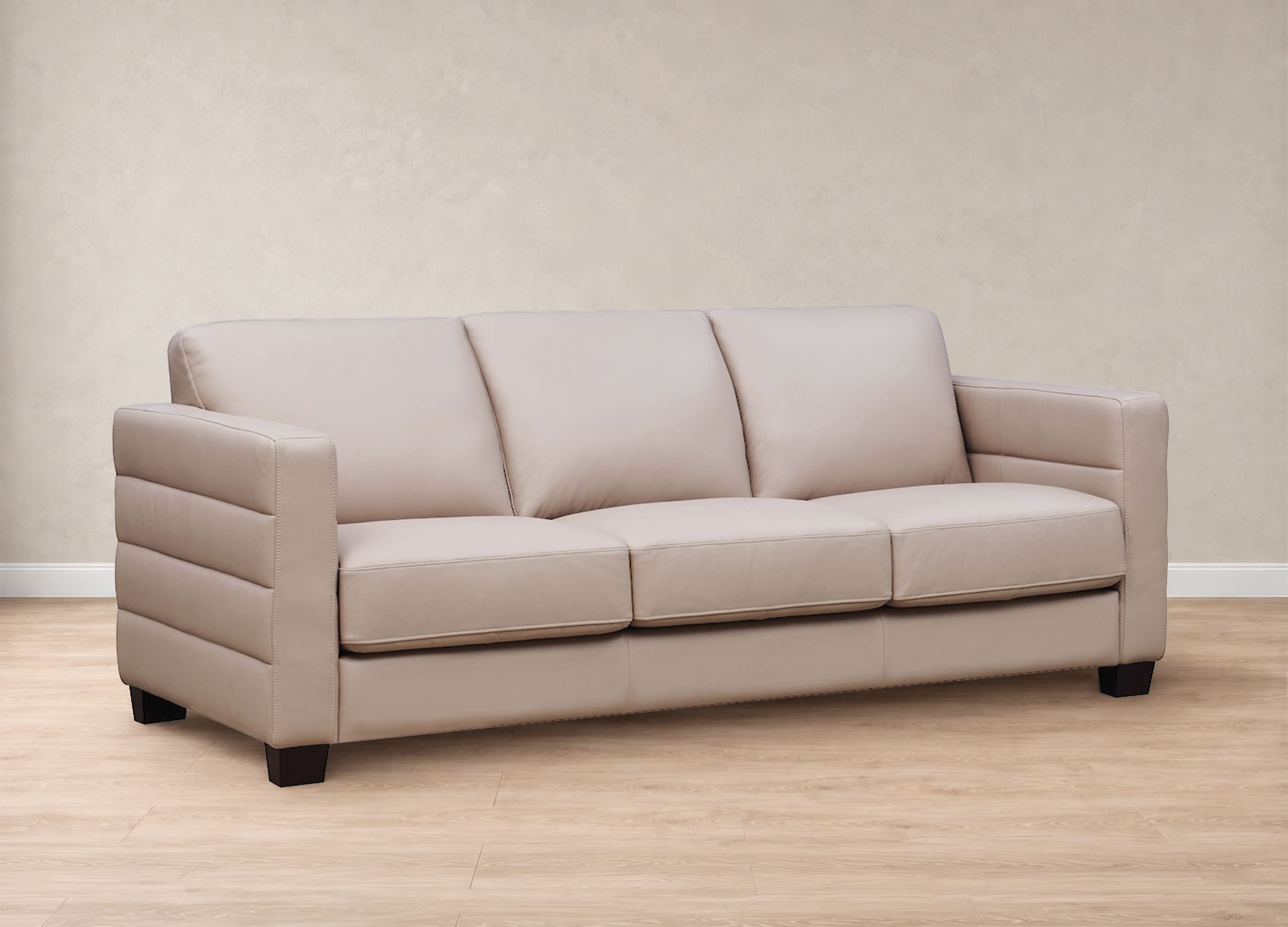 A three-seat upholstered sofa in a light beige or taupe leather-like material, shown in a staged room with a textured beige wall and light wood floor.