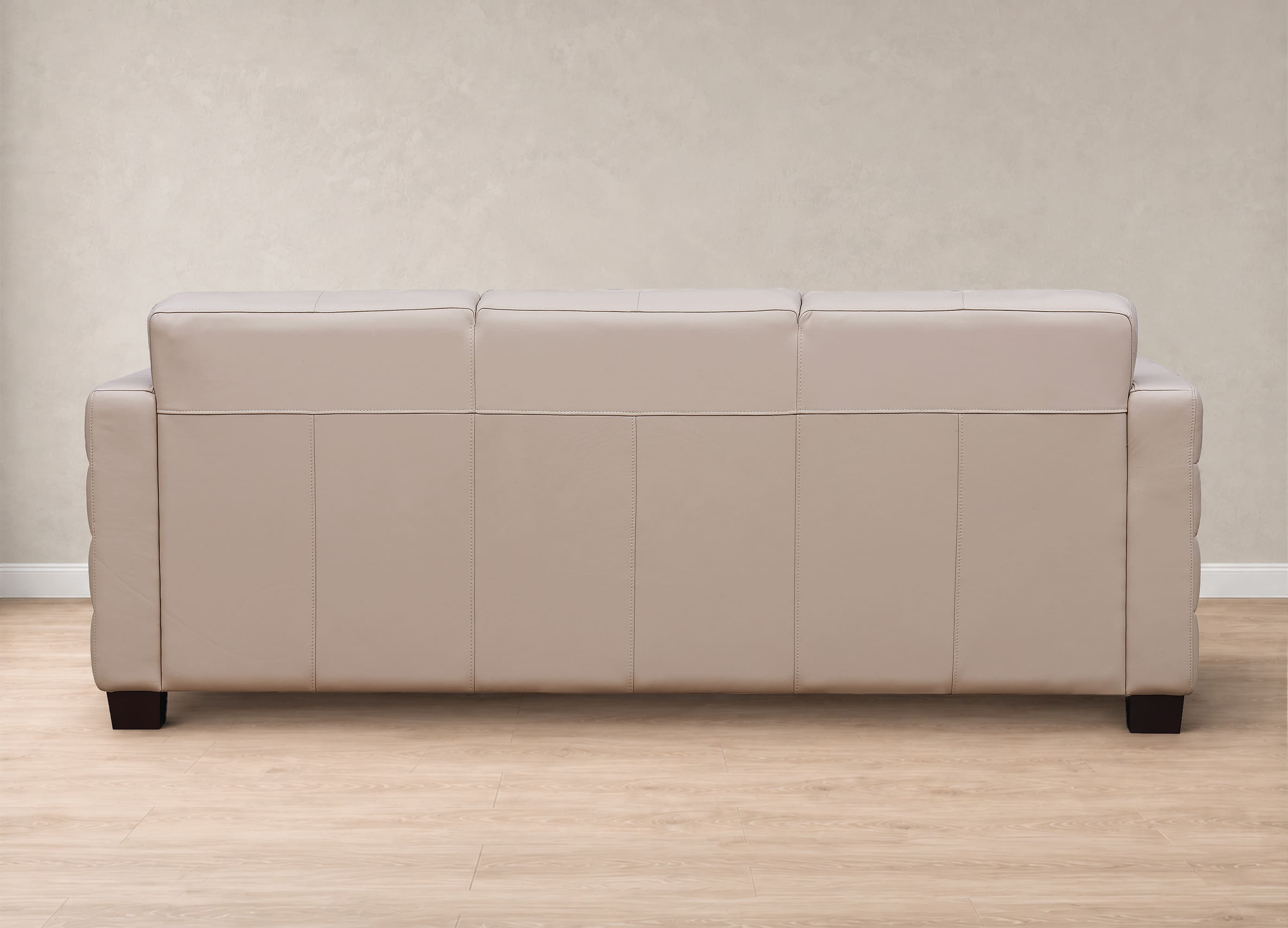 A three-seat sofa upholstered in light beige leather, shown from the back against a textured beige wall and light wood floor.