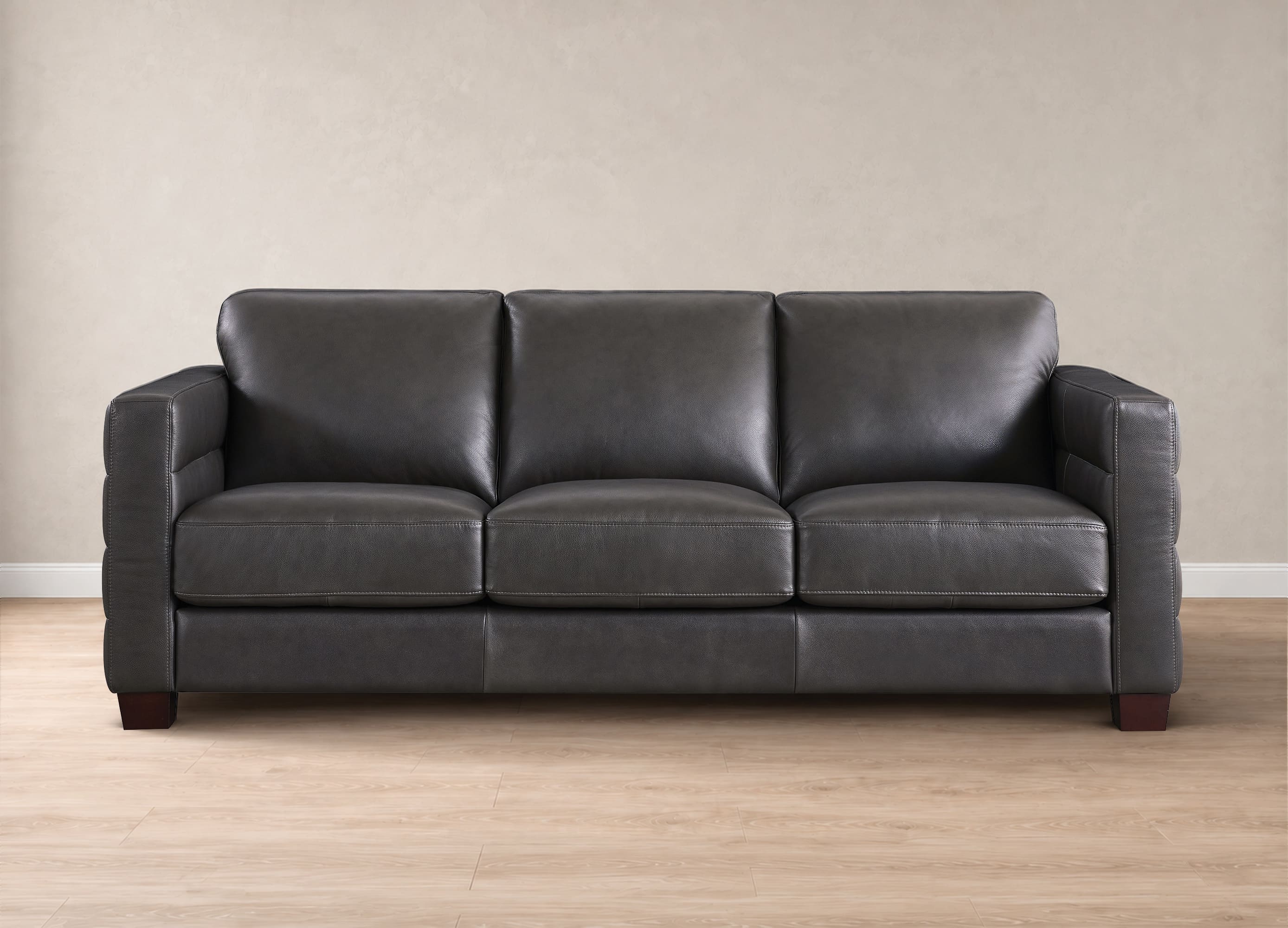 A three-seat dark gray leather sofa with a modern low-profile design, set against a neutral beige wall and light wood floor.