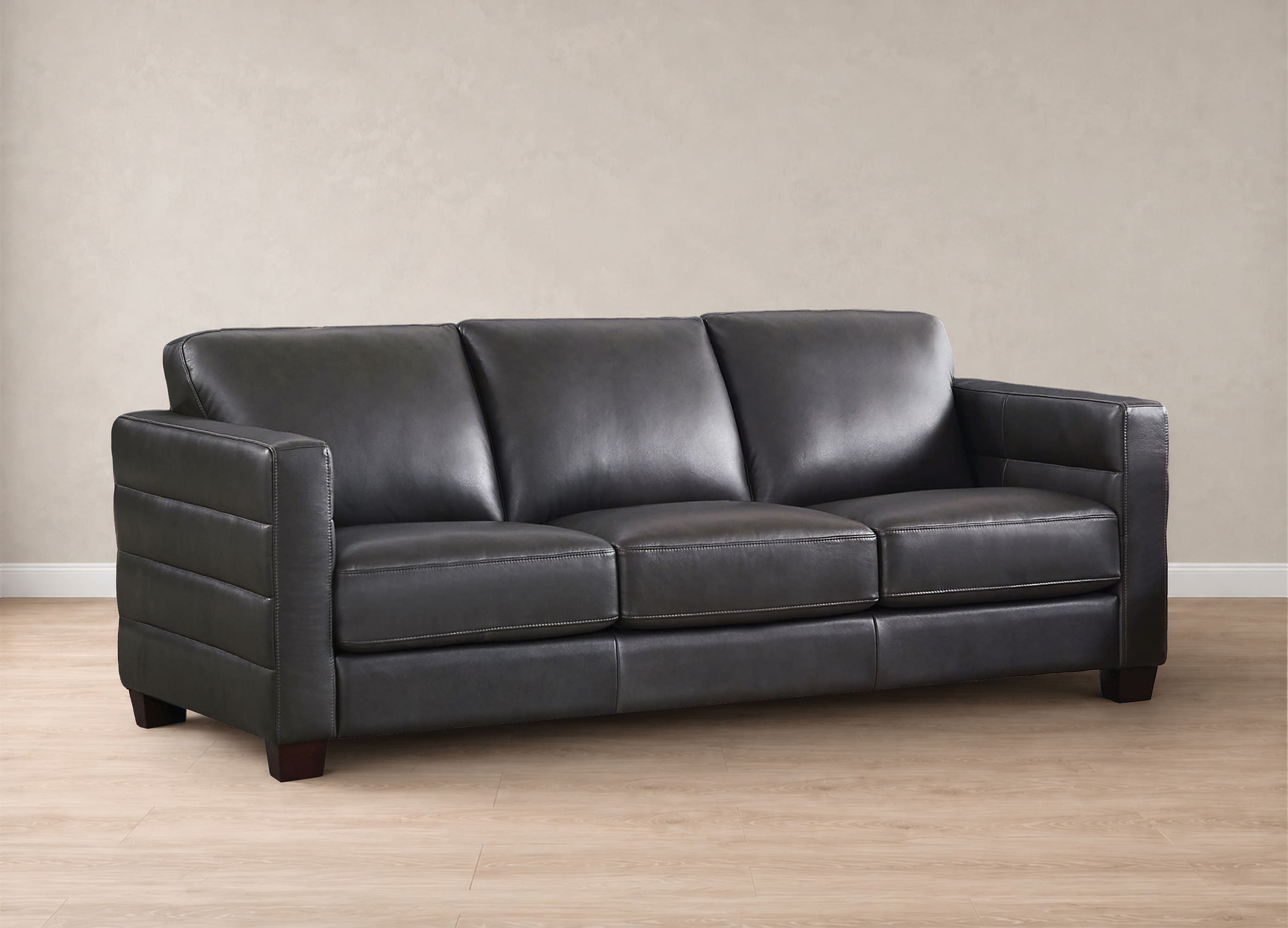 A three-seat black leather sofa with tufted armrests, shown in a studio setting with a beige textured wall and light wood floor.