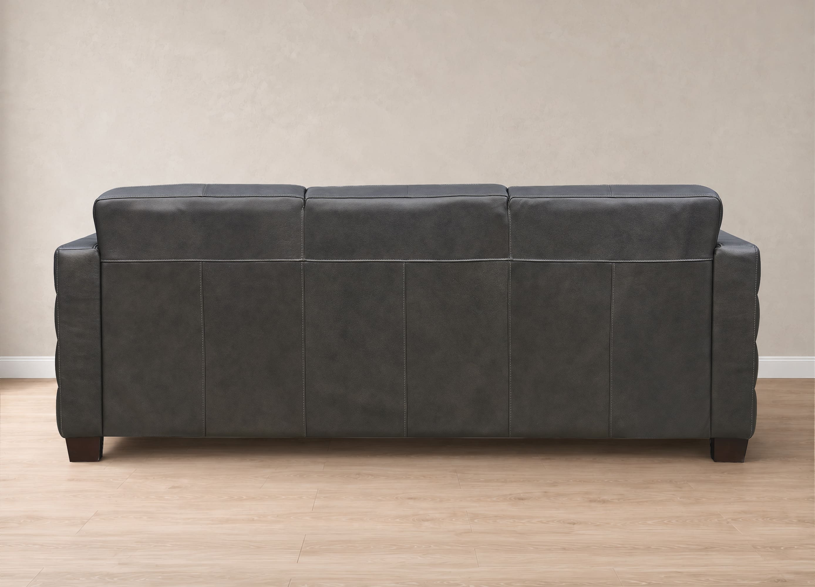 A three-seat sofa upholstered in dark charcoal gray faux leather, shown from the back view against a neutral beige wall and light wood floor.