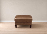 A brown leather ottoman with dark wooden legs, positioned centrally on a light wood floor against a textured beige wall.