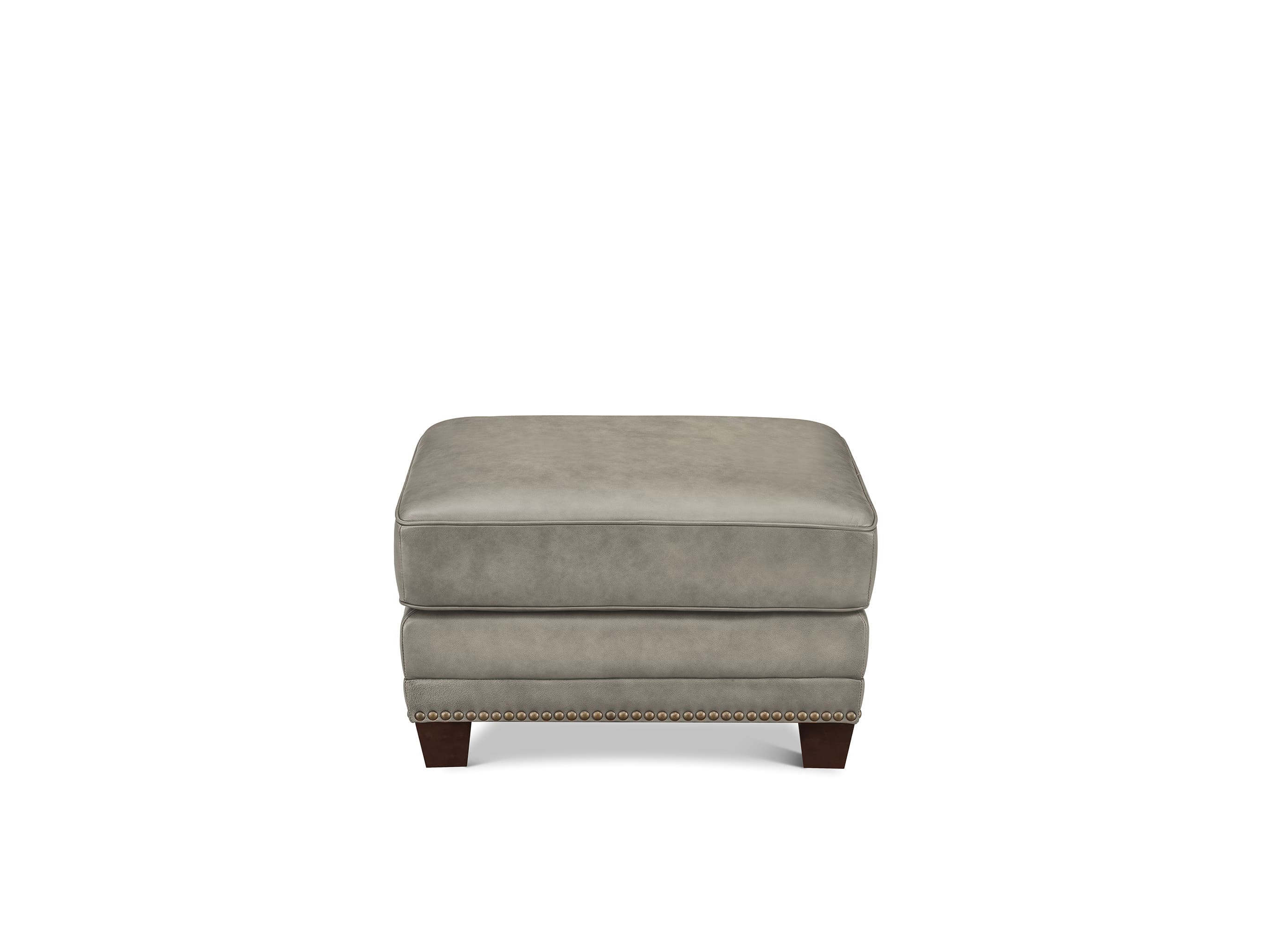 A square upholstered ottoman in a light taupe-gray fabric, featuring dark brown wooden legs and nailhead trim along the front edge, shown against a plain white background.