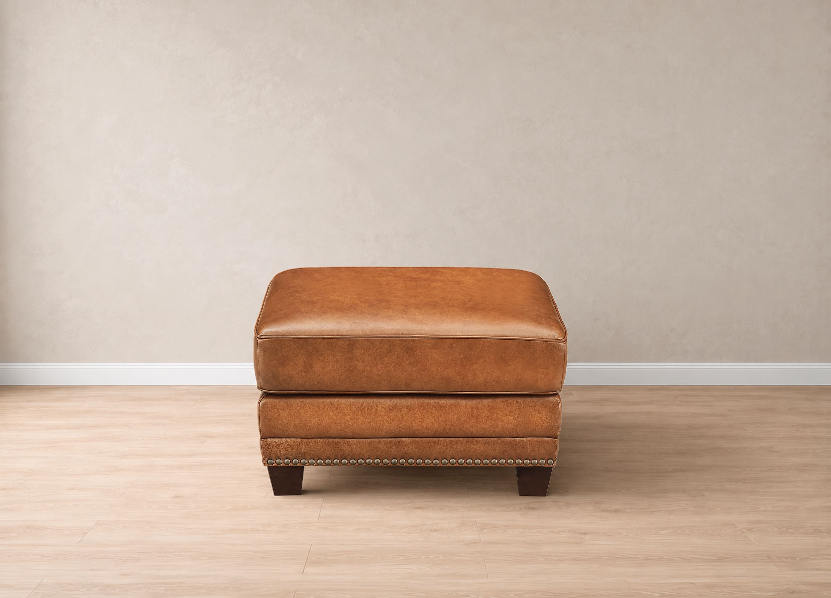 A square brown leather ottoman with dark wooden legs and nailhead trim, centered against a neutral beige wall and light wood floor.